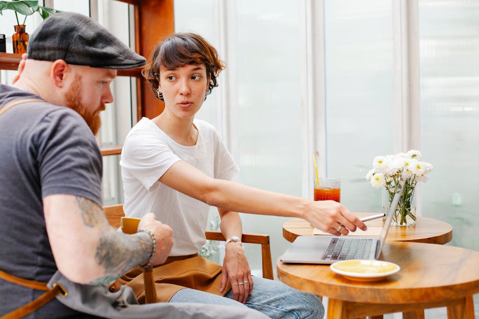 Young female barista pointing at netbook screen while talking to bearded male coworker in cafeteria
