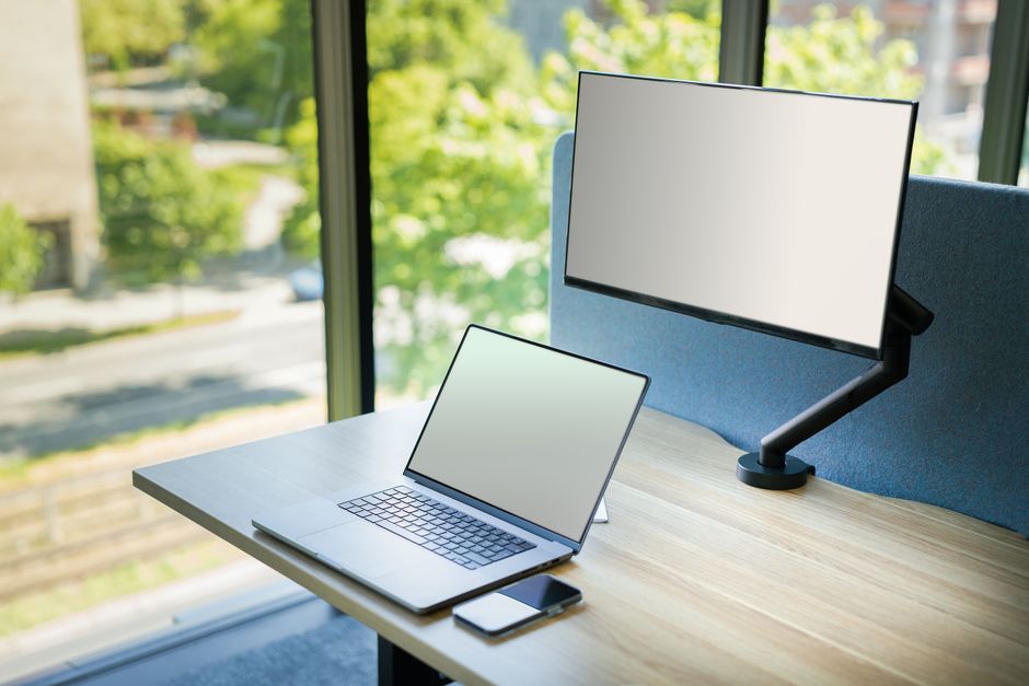 A contemporary office desk setup with a laptop and monitor by a window, featuring bright natural light.