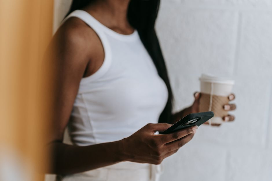 A woman in a white tank top holding a smartphone and a coffee cup indoors.