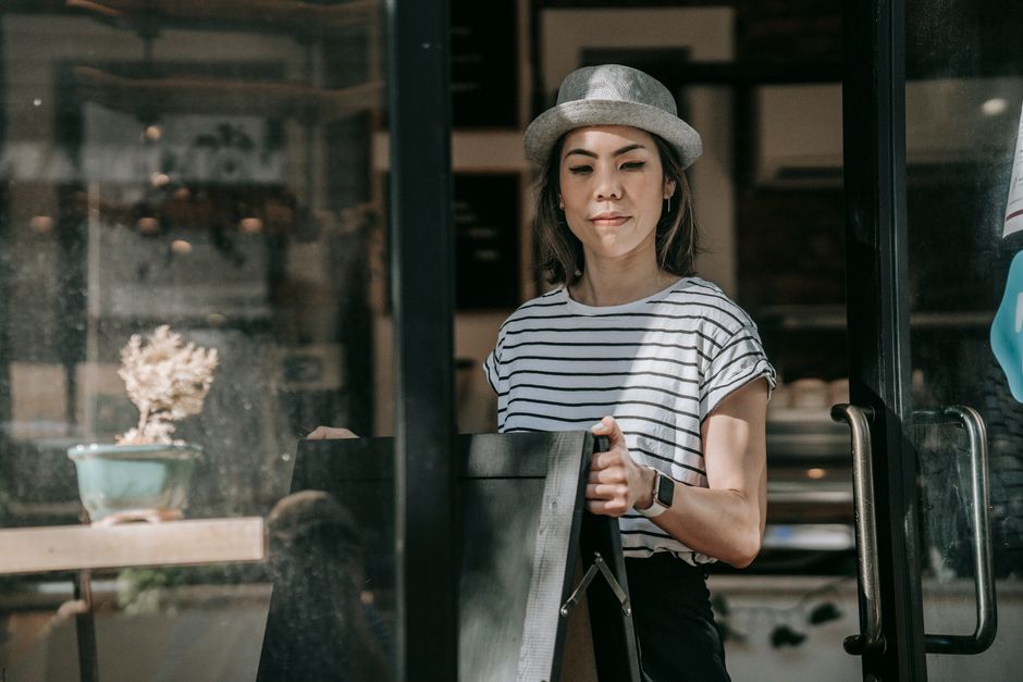 Woman in casual attire positioning a sign outside a cafe, wearing a hat. Bright daylight, modern aesthetic.