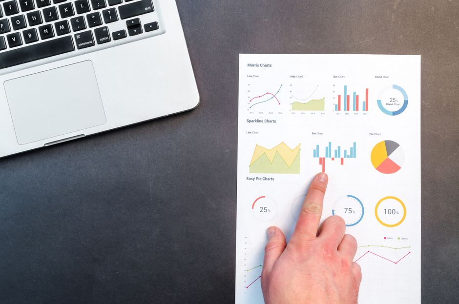 A hand pointing at colorful charts with a laptop in the background on a desk.