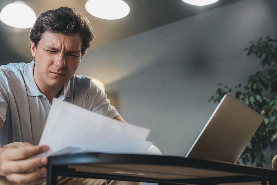 Man focusing intently while reviewing documents at a desk with a laptop.